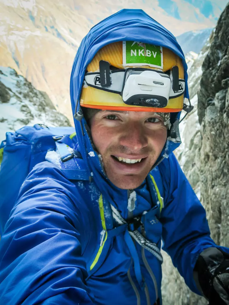 Een bergbeklimmer met een blauwe jas, helm en hoofdlamp glimlacht terwijl hij een besneeuwde, rotsachtige berg beklimt.